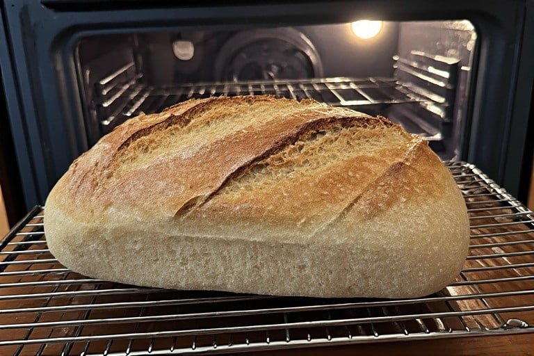 Overhead shot of a sourdough loaf cooling correctly on a wire rack, oven door slightly open in background, natural kitchen setting, warm light