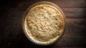 A glass bowl on a dark kitchen counter at night showing sourdough dough with visible bubbles along the edges, soft ambient lighting, overhead shot, rustic wood surface